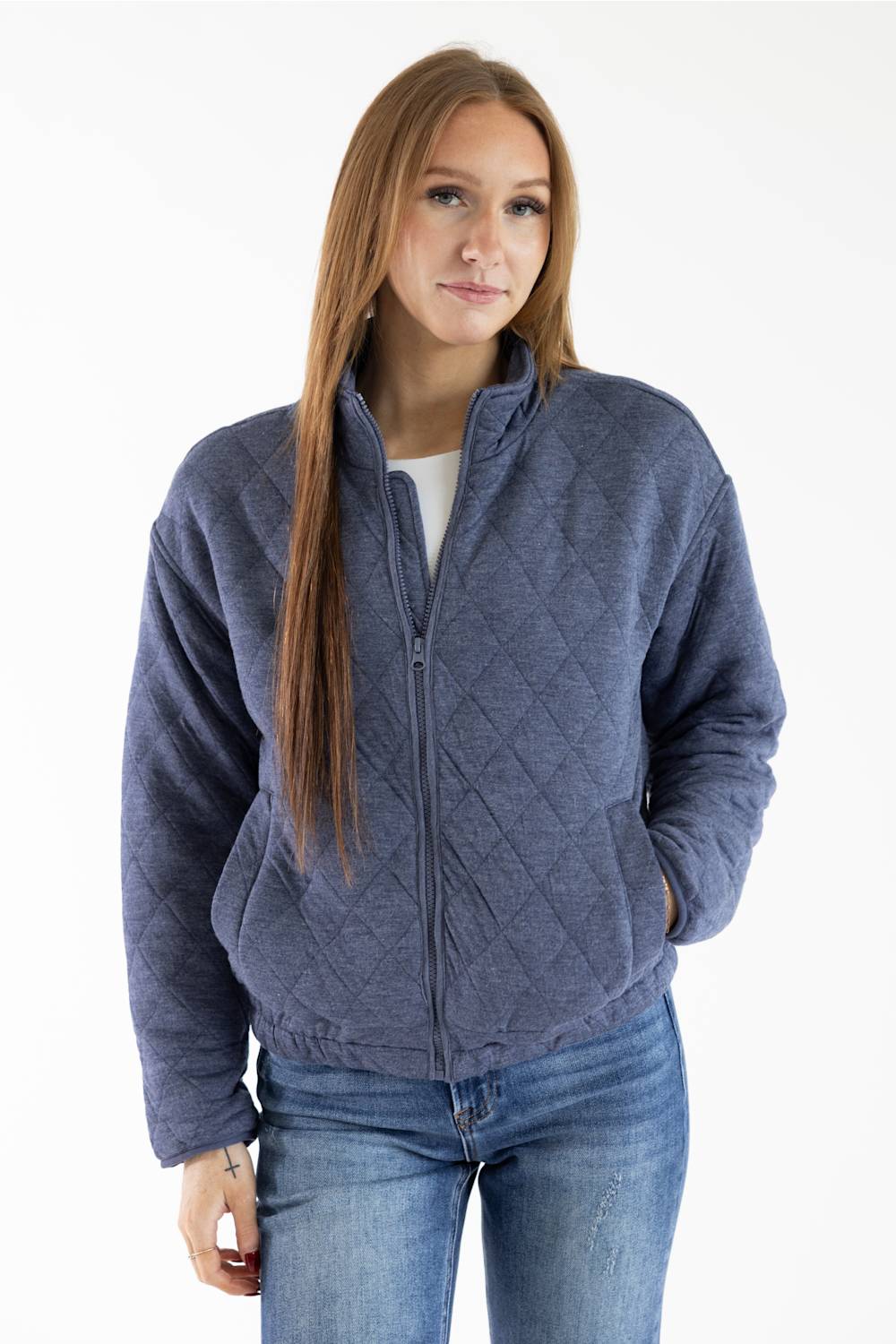 A woman modeling a slate blue diamond-quilted zip-up fleece jacket, paired with a white top and blue jeans, against a plain white studio background.