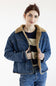 A woman wearing a blue denim jacket with a tan shearling-style collar and lining, layered over a striped sweater and blue jeans, against a plain white studio background.