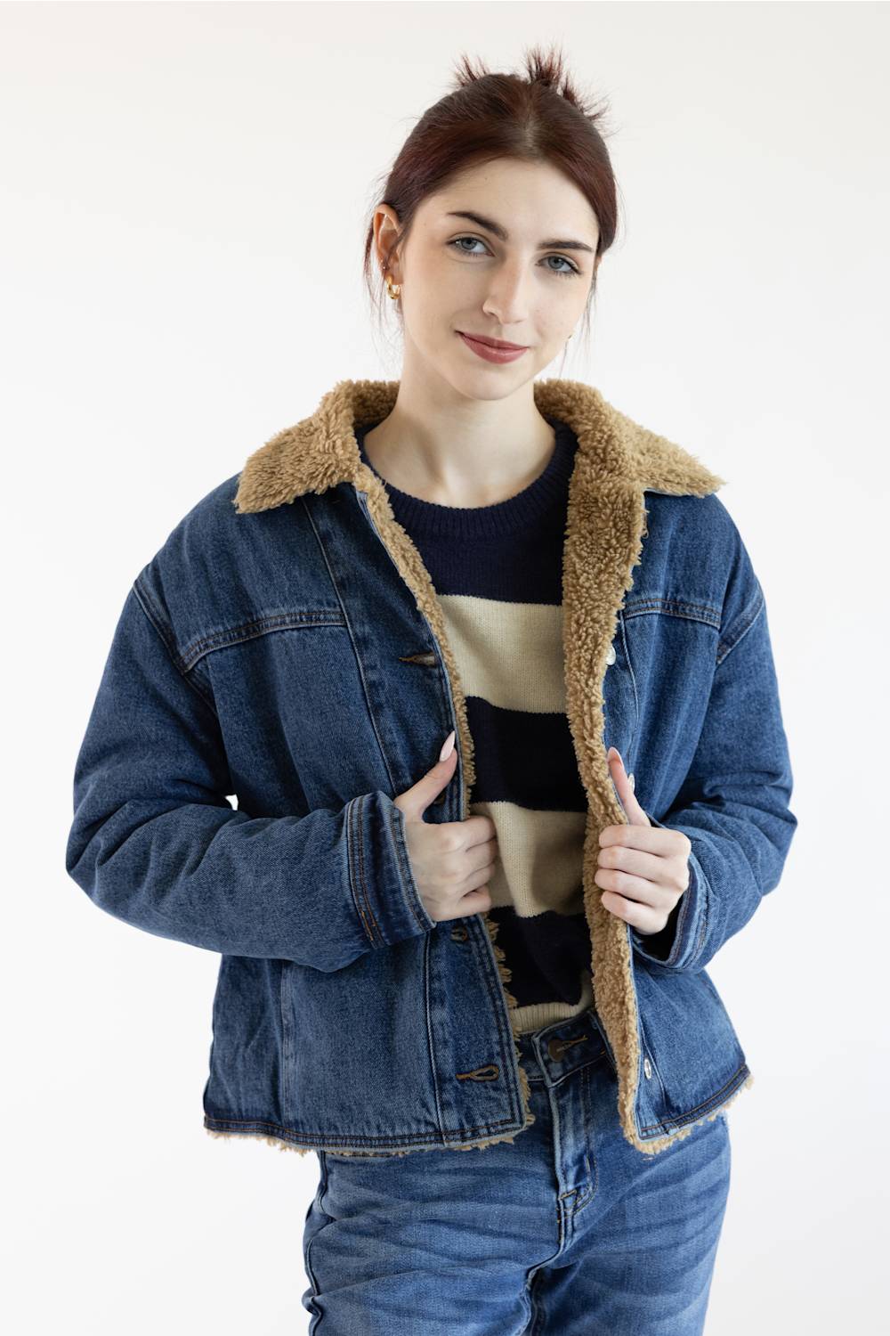 A woman wearing a blue denim jacket with a tan shearling-style collar and lining, layered over a striped sweater and blue jeans, against a plain white studio background.