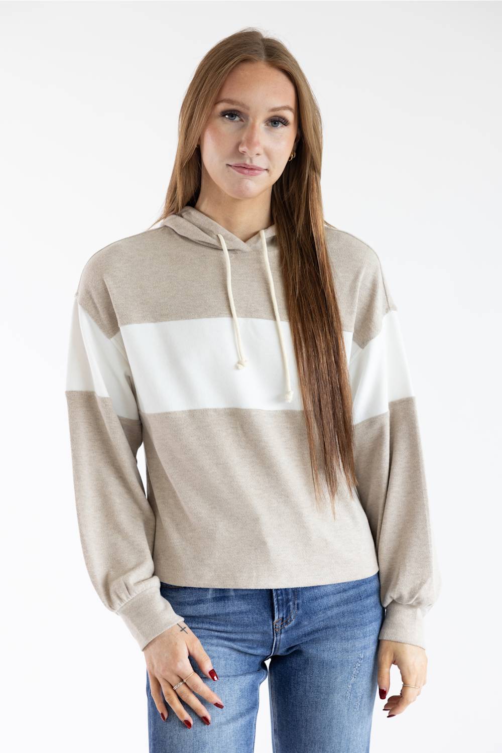 A woman modeling a color-blocked knit hoodie with beige, white, and light brown horizontal stripes, paired with blue jeans, against a plain white studio background.