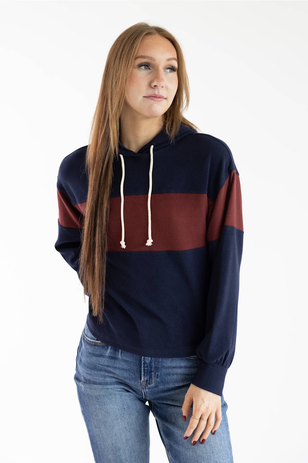 A woman modeling a colorblock hoodie with navy blue, burgundy, and rust orange horizontal stripes, paired with blue jeans, against a plain white studio background.