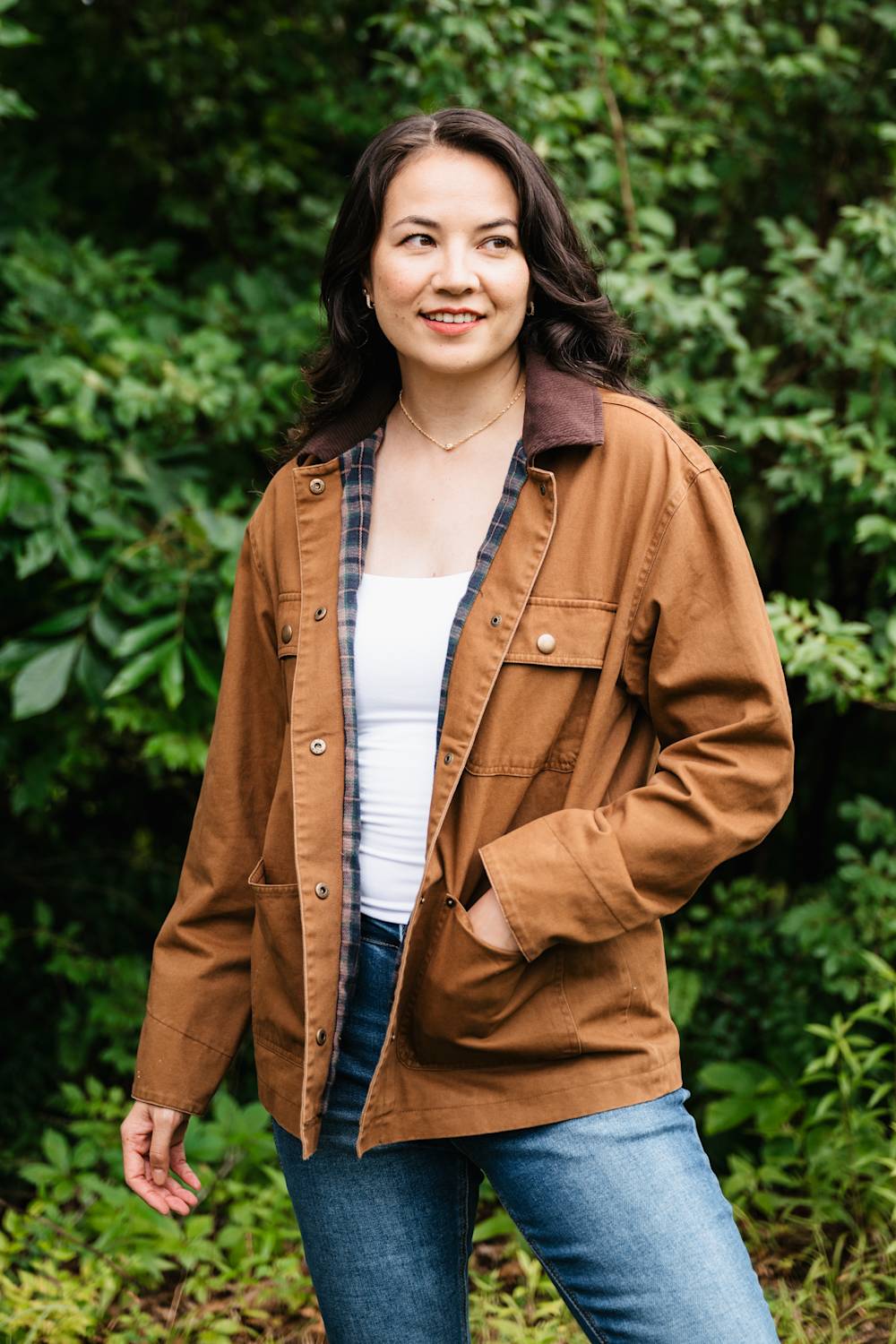 A woman wearing a brown utility jacket with a dark brown collar, layered over a plaid shirt and white top, paired with blue jeans, standing in front of a lush green forest background.