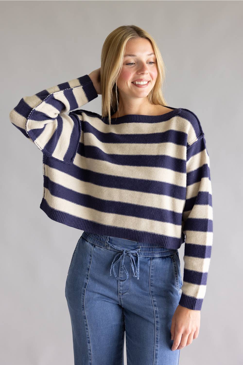 A woman wearing a cropped knit sweater with horizontal navy and cream stripes, paired with blue jeans, standing against a plain gray studio background.
