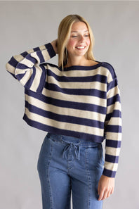 A woman wearing a cropped knit sweater with horizontal navy and cream stripes, paired with blue jeans, standing against a plain gray studio background.
