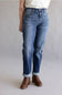 A person wearing medium-wash blue jeans with rolled cuffs and brown ankle boots, shown from the waist down against a light gray studio background.