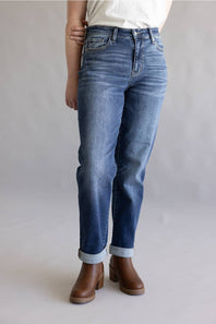 A person wearing medium-wash blue jeans with rolled cuffs and brown ankle boots, shown from the waist down against a light gray studio background.