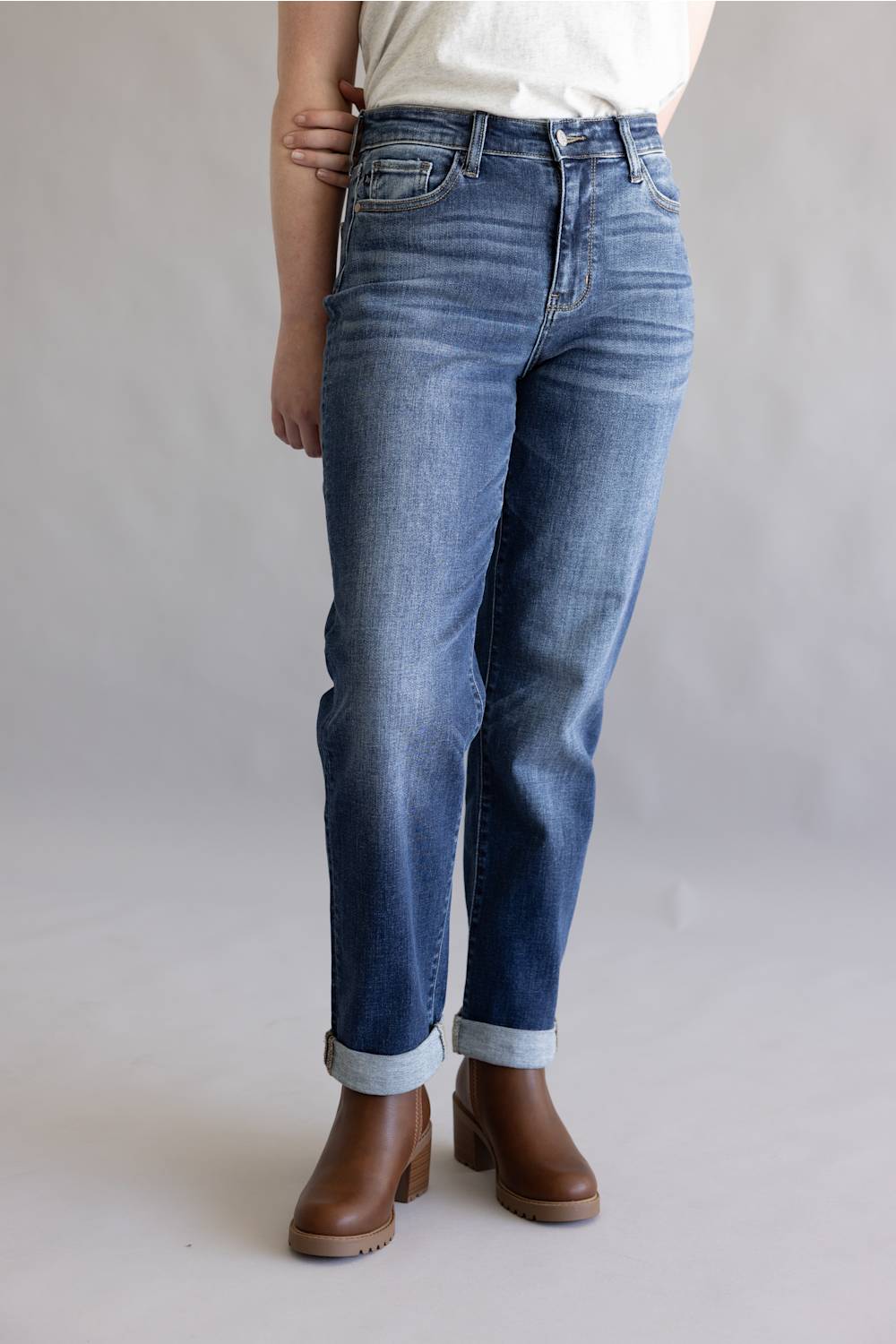 A person wearing medium-wash blue jeans with rolled cuffs and brown ankle boots, shown from the waist down against a light gray studio background.
