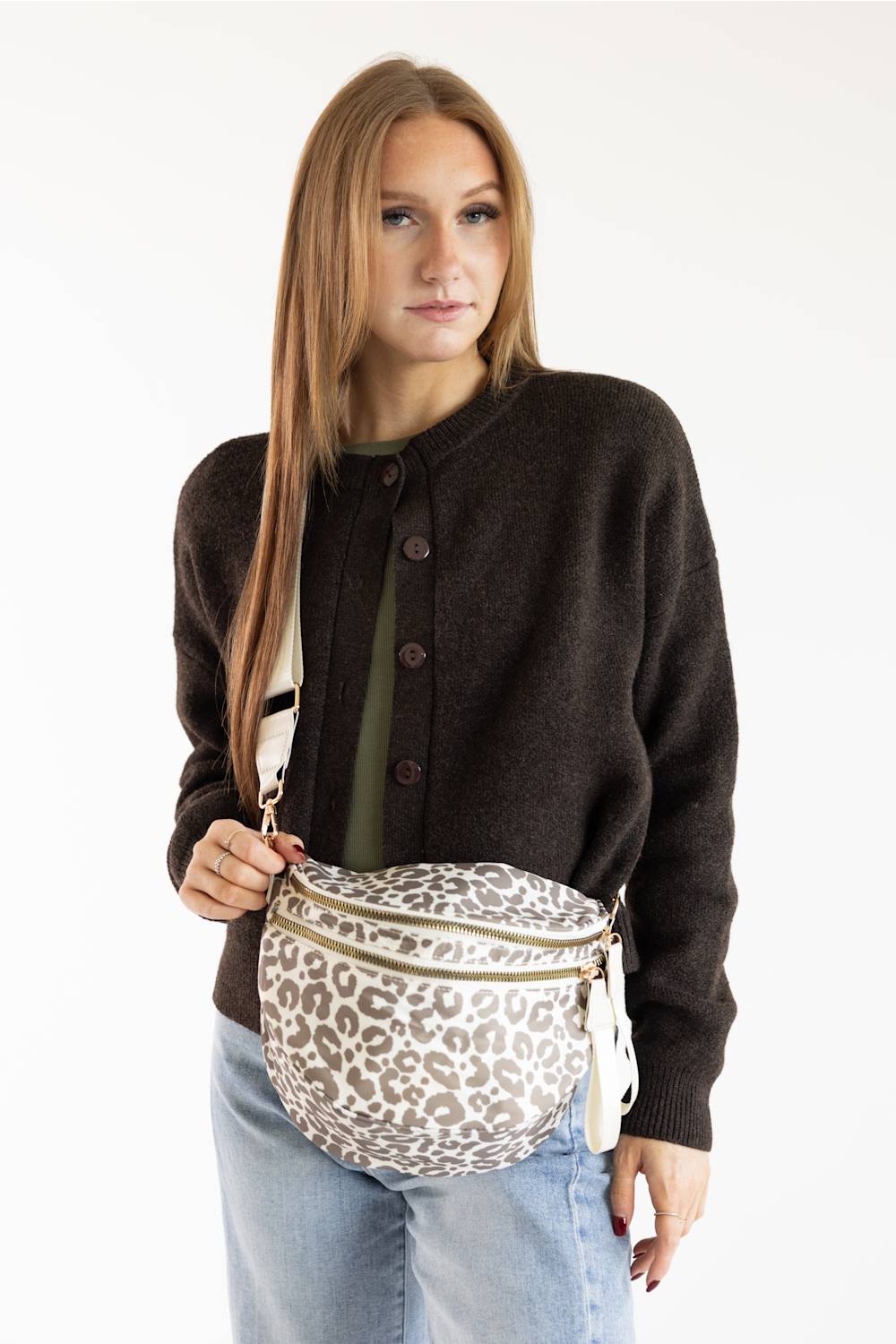 A woman wearing a dark brown button-up cardigan, olive green shirt, and light blue jeans, holding a leopard print fanny pack against a plain white background.