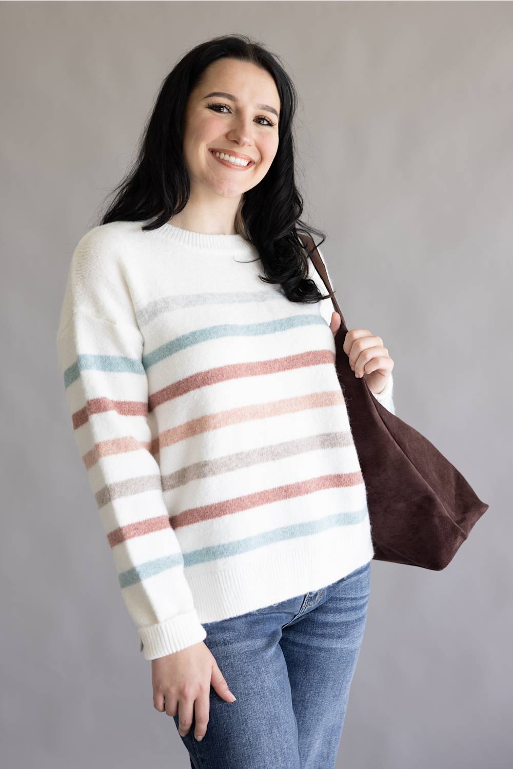 A woman wearing a white knit sweater with horizontal dusty pink and light blue stripes, paired with blue jeans and carrying a brown tote bag, set against a plain gray studio background.
