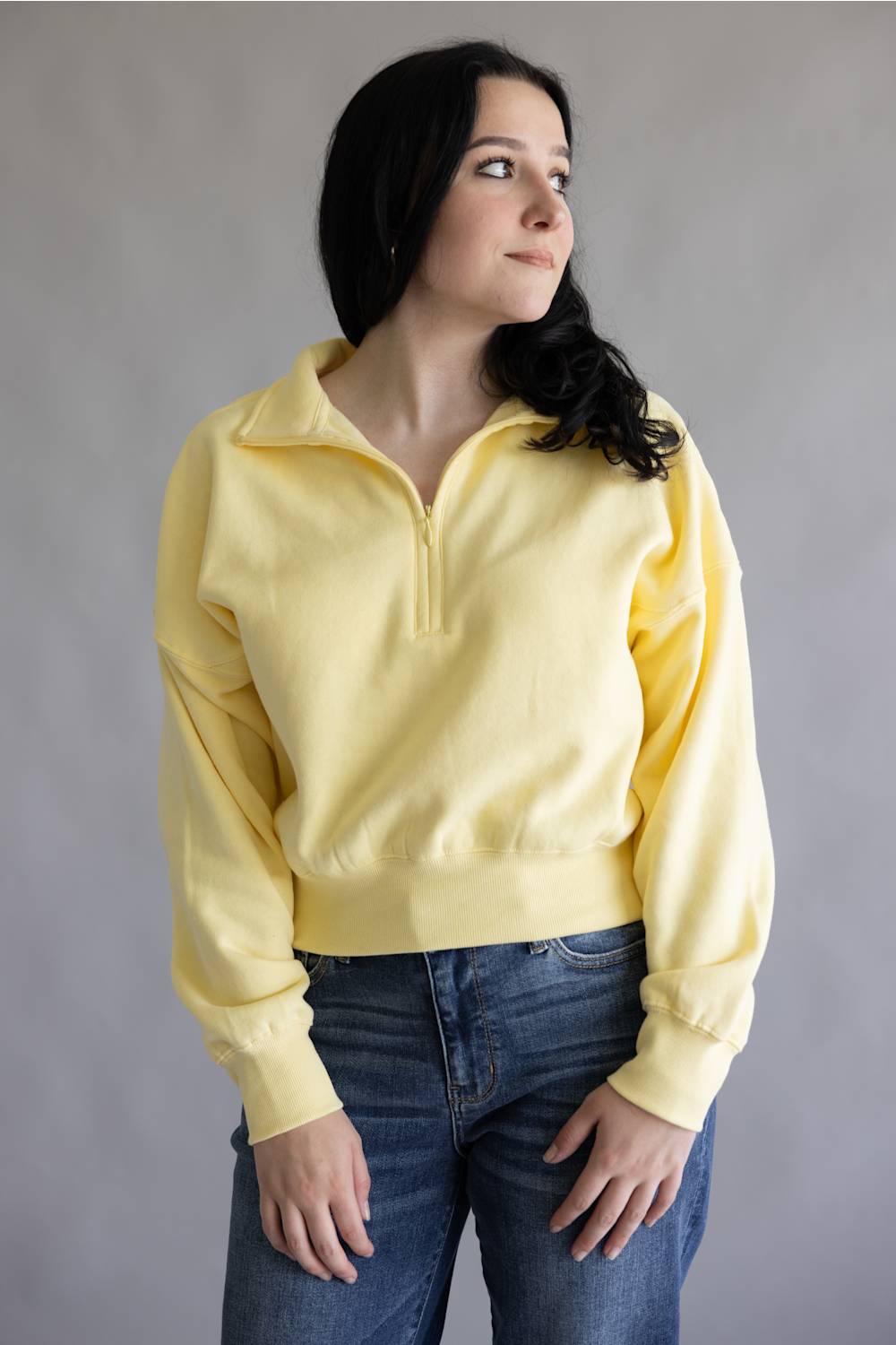 A woman wearing a pale yellow half-zip sweatshirt and blue jeans, standing against a plain gray studio background.