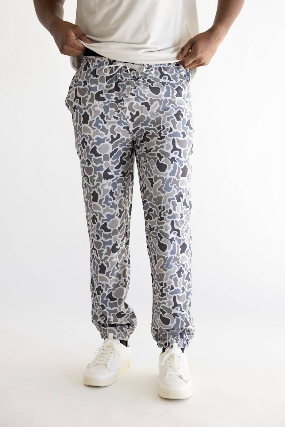 A person wearing loose-fit jogger-style pants with an allover abstract camouflage-like print in gray, white, and black, paired with a white T-shirt and white sneakers against a plain light background.