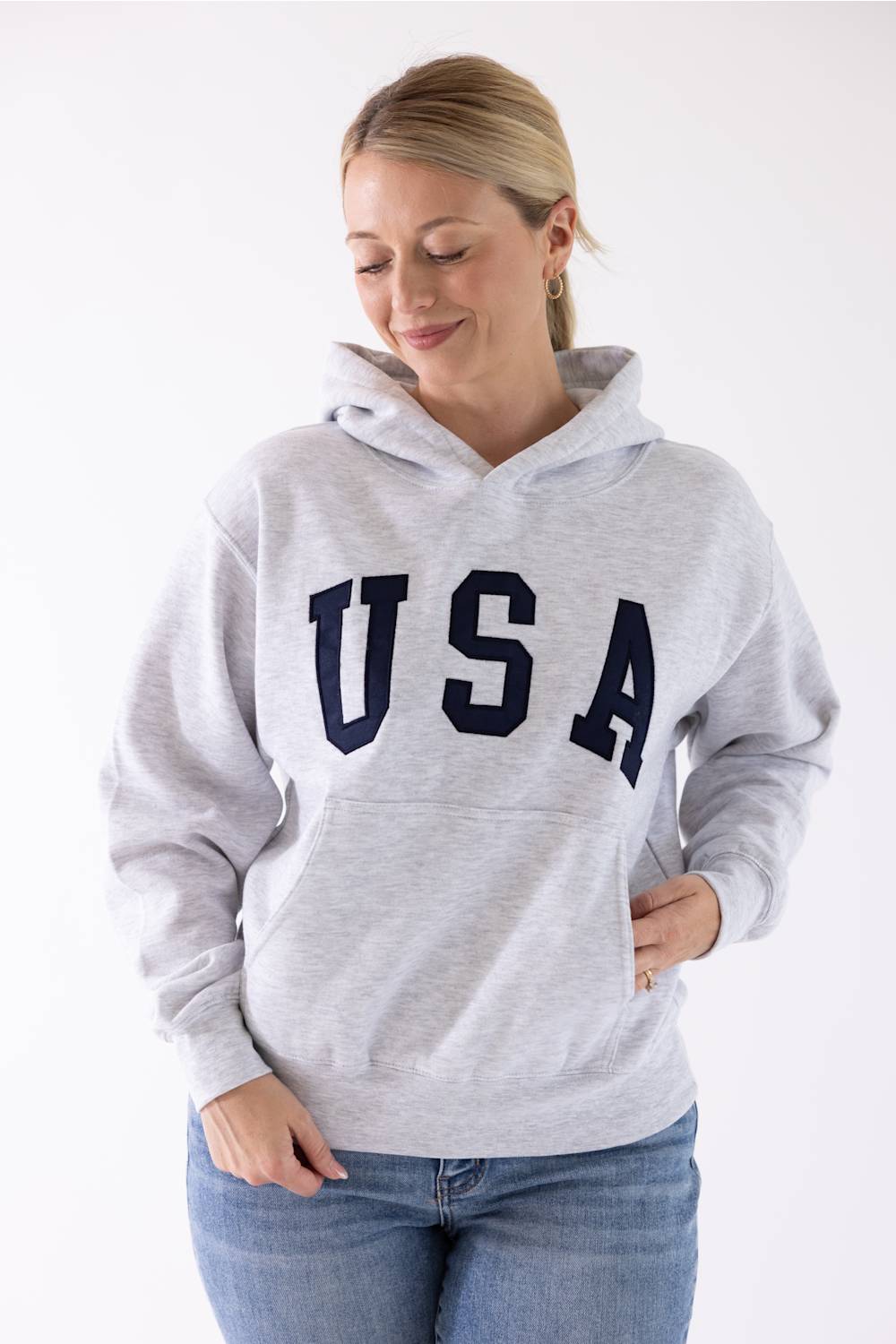 Front view of a blonde woman wearing an ice grey Reflex fleece pullover hoodie featuring large dark navy "USA" block lettering across the chest and a front kangaroo pocket.