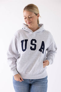 Front view of a blonde woman wearing an ice grey Reflex fleece pullover hoodie featuring large dark navy "USA" block lettering across the chest and a front kangaroo pocket.