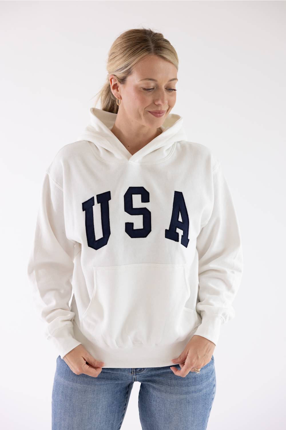 Front view of a blonde woman wearing an ivory Reflex fleece pullover hoodie, blue jeans, and gold hoop earrings.