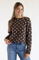 Front view of a smiling model wearing the Just Polly dark brown long-sleeve knit sweater featuring an ivory polka dot pattern, a slightly rolled boat neckline, and ribbed cuffs, styled with denim jeans against a plain light background.