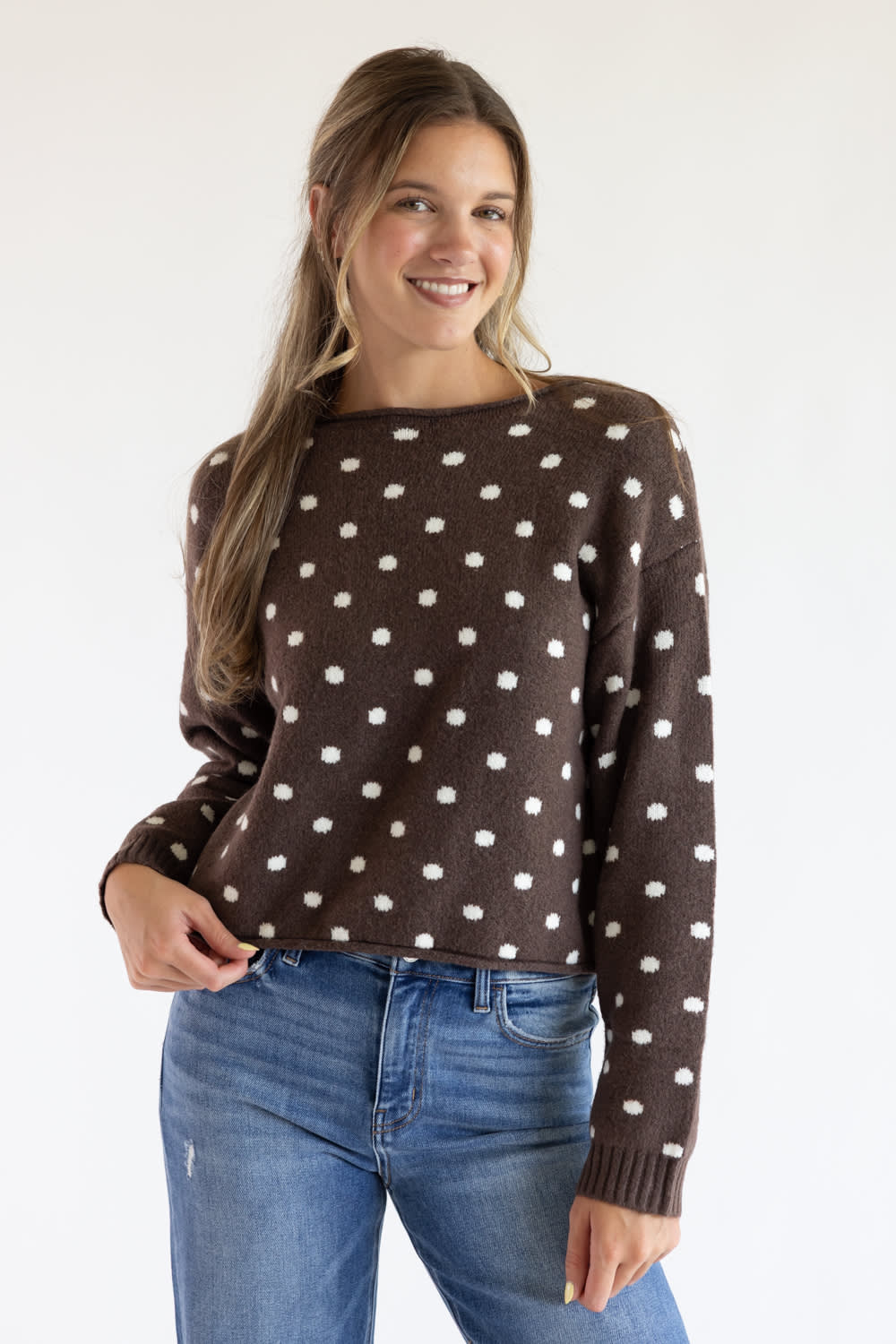 Front view of a smiling model wearing the Just Polly dark brown long-sleeve knit sweater featuring an ivory polka dot pattern, a slightly rolled boat neckline, and ribbed cuffs, styled with denim jeans against a plain light background.