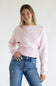 Front view of a smiling model wearing the Just Polly light pink long-sleeve knit sweater featuring a white polka dot pattern, a boat neckline, and wide ribbed hem and cuffs, styled with denim jeans against a plain light background.