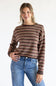 Front view of a smiling model posing with her hand in her pocket, wearing the Just Polly long-sleeve knit sweater featuring multi-color horizontal stripes in brown, red, and tan, a slightly rolled boat neckline, and ribbed cuffs, styled with denim jeans against a plain white background.