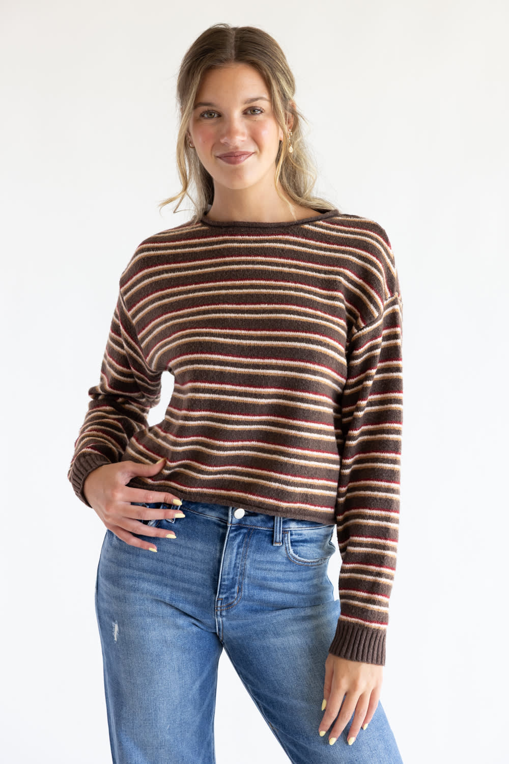 Front view of a smiling model posing with her hand in her pocket, wearing the Just Polly long-sleeve knit sweater featuring multi-color horizontal stripes in brown, red, and tan, a slightly rolled boat neckline, and ribbed cuffs, styled with denim jeans against a plain white background.