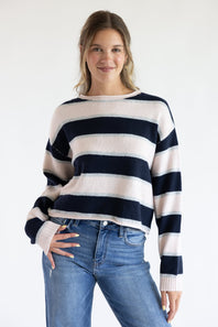 Front view of a model wearing the Just Polly long-sleeve knit sweater featuring wide light pink and navy blue horizontal stripes accented with thin heather grey stripes, a boat neckline, and ribbed cuffs, styled with denim jeans against a plain white background.