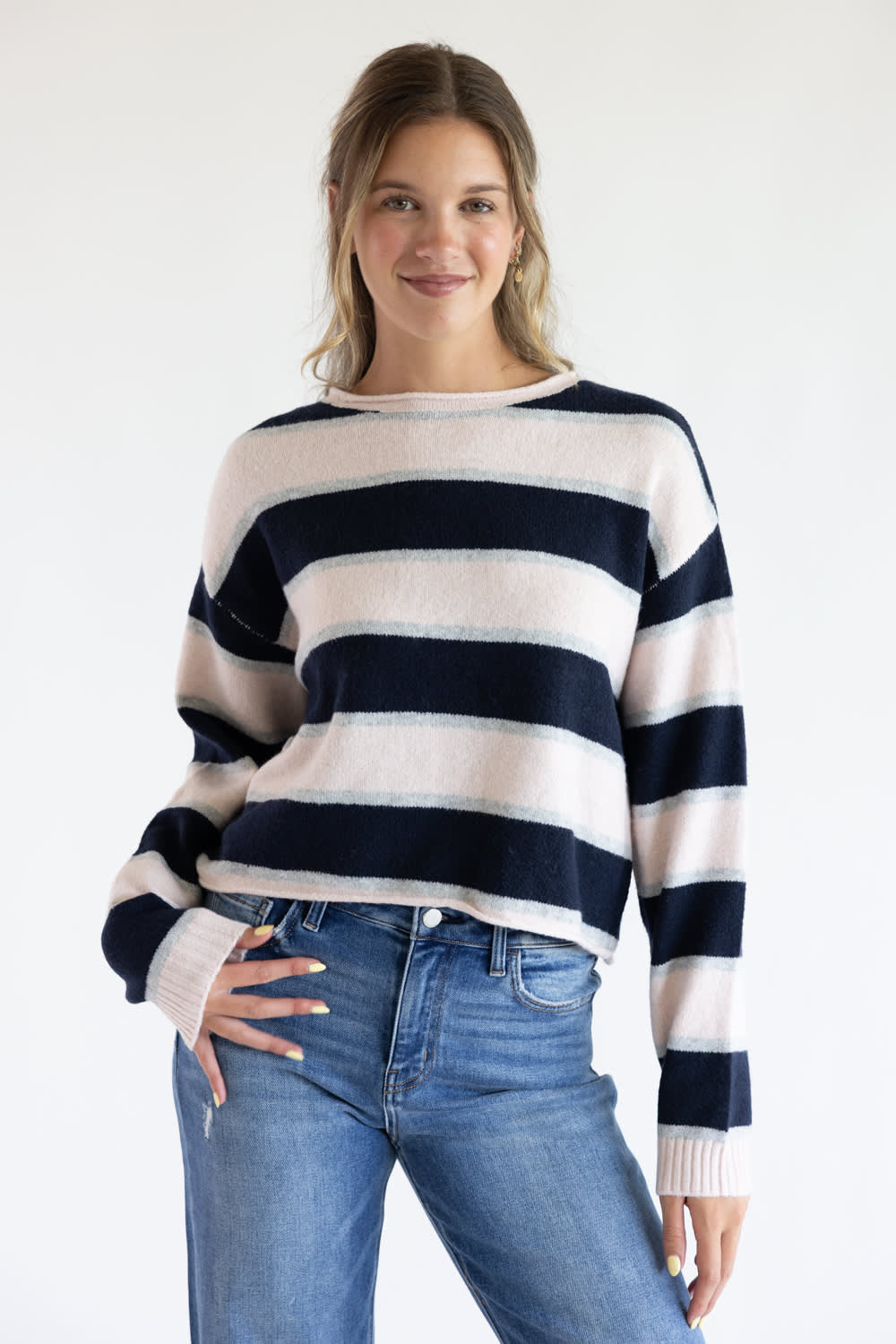Front view of a model wearing the Just Polly long-sleeve knit sweater featuring wide light pink and navy blue horizontal stripes accented with thin heather grey stripes, a boat neckline, and ribbed cuffs, styled with denim jeans against a plain white background.
