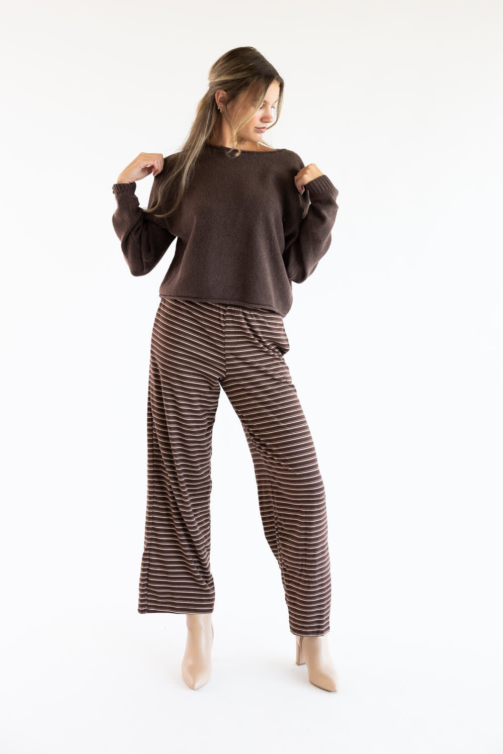Model wearing the Just Polly dark brown boat neck sweater featuring ribbed cuffs, styled with brown and white striped wide-leg trousers and beige boots against a white studio background.