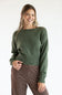 Front view of a smiling model wearing the Just Polly olive green long-sleeve knit sweater featuring a slightly rolled boat neckline and wide ribbed hem and cuffs, styled with brown striped pants against a plain white background.