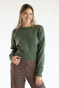 Front view of a smiling model wearing the Just Polly olive green long-sleeve knit sweater featuring a slightly rolled boat neckline and wide ribbed hem and cuffs, styled with brown striped pants against a plain white background.