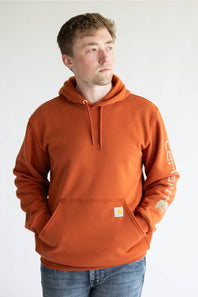 Man wearing a burnt orange colored hoodie, with Carhartt logo down the left arm sleeve. 
