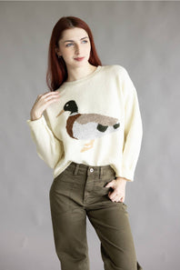 A woman with red hair wearing a cream-colored knit sweater with a detailed duck motif on the front, paired with olive green pants, standing against a neutral gray studio background.