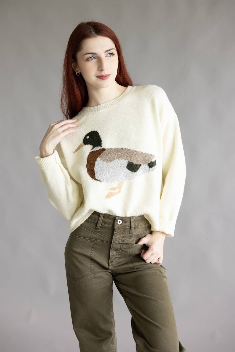 A woman with red hair wearing a cream-colored knit sweater with a detailed duck motif on the front, paired with olive green pants, standing against a neutral gray studio background.