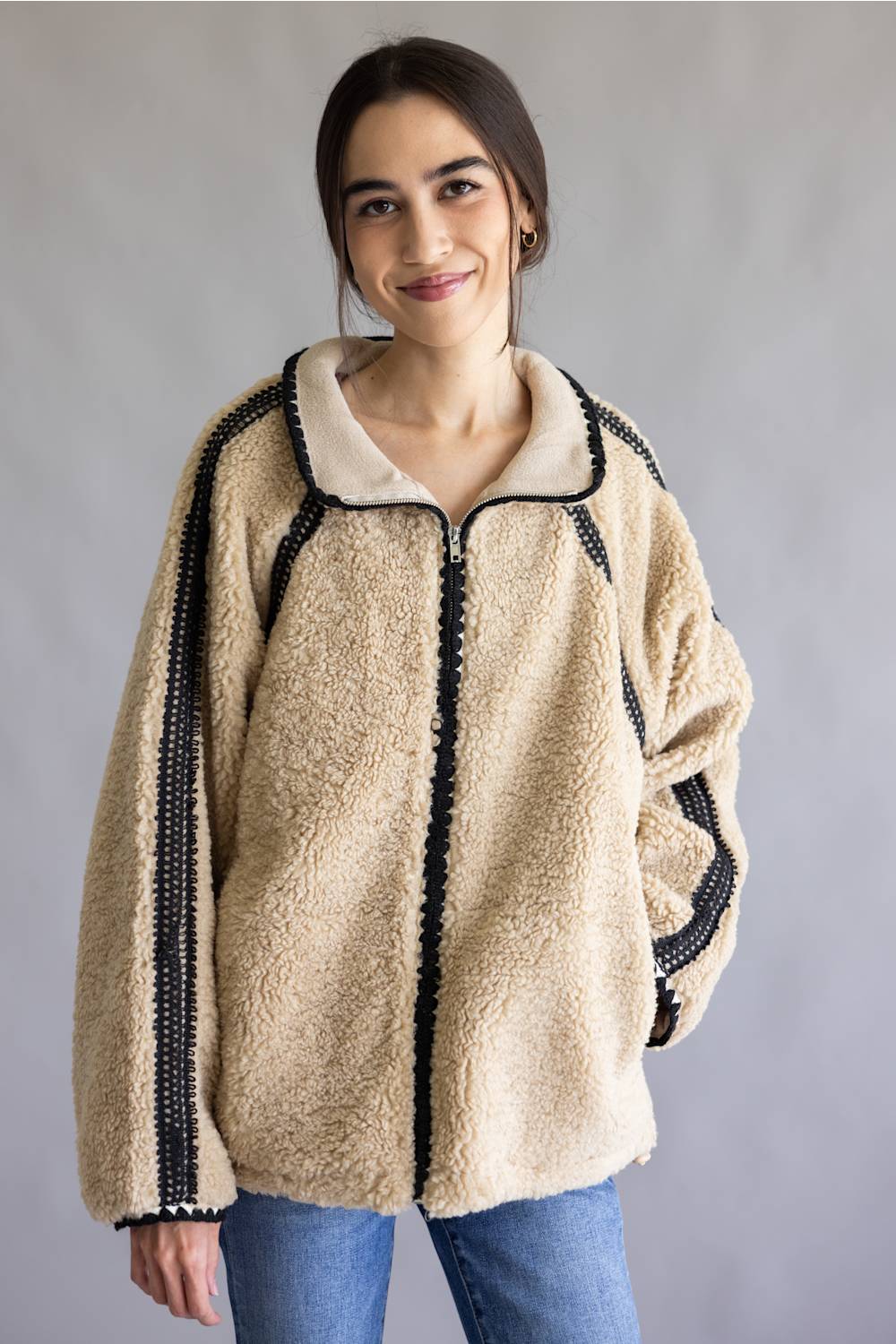 A woman wearing a beige sherpa fleece zip-up jacket with black beaded trim down the front and along the sleeves, paired with blue jeans, set against a plain gray studio background.