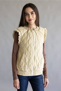 A young girl modeling a cream-colored diamond-quilted sleeveless vest with ruffled shoulders, worn over a brown long-sleeve shirt and blue jeans, against a plain gray studio background.