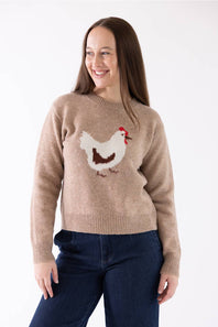 A woman wearing a beige crewneck sweater with a white, brown, and red chicken motif on the front, paired with dark blue jeans, against a plain white background.