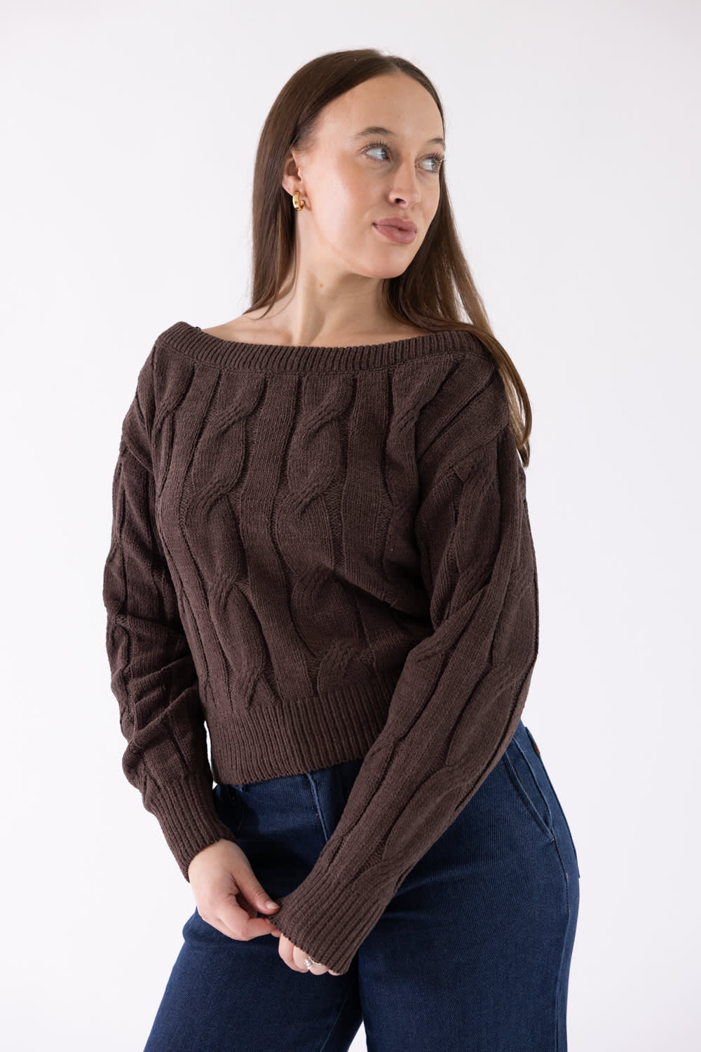 A woman wearing a dark brown cable-knit off-the-shoulder sweater and dark blue jeans, standing against a plain white studio background.