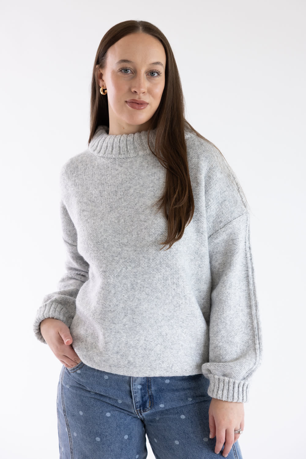 A woman wearing a light gray heathered knit sweater with a mock neck and ribbed cuffs, paired with blue jeans, photographed against a plain white studio background.