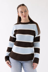 A woman wearing a knit sweater with horizontal stripes in light blue, off-white, and dark brown, paired with light-wash jeans, against a plain white studio background.