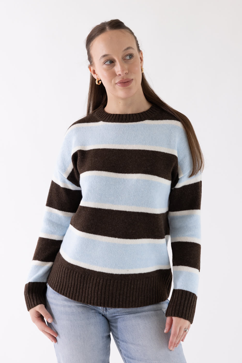 A woman wearing a knit sweater with horizontal stripes in light blue, off-white, and dark brown, paired with light-wash jeans, against a plain white studio background.