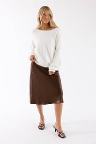 A woman modeling a white knit sweater and a brown midi skirt against a plain white studio background.