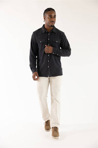 A man modeling a black long-sleeve button-up shirt with chest pockets, paired with off-white trousers and brown shoes, against a plain white studio background.