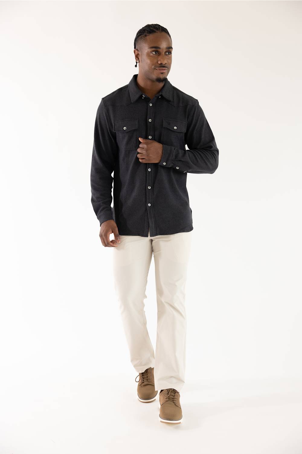 A man modeling a black long-sleeve button-up shirt with chest pockets, paired with off-white trousers and brown shoes, against a plain white studio background.
