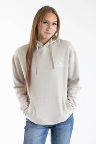 A woman with long light brown hair wearing a light beige pullover hoodie with a small white graphic on the left chest, paired with blue jeans, against a plain white background.