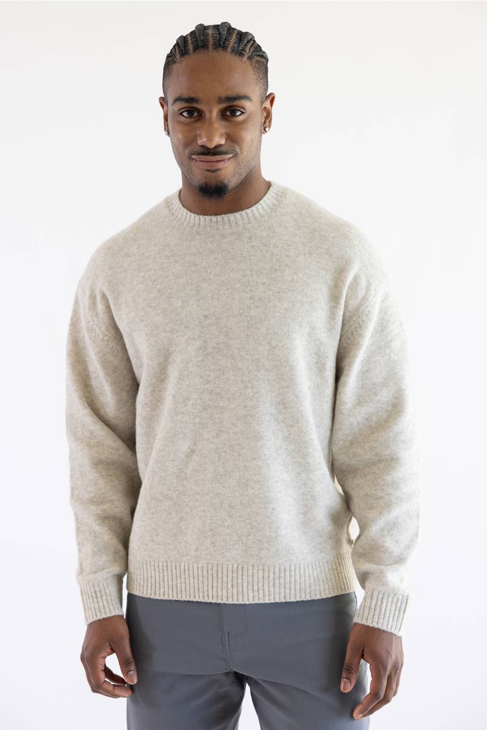 A man wearing a light beige knit crewneck sweater and gray trousers, standing against a plain white studio background.