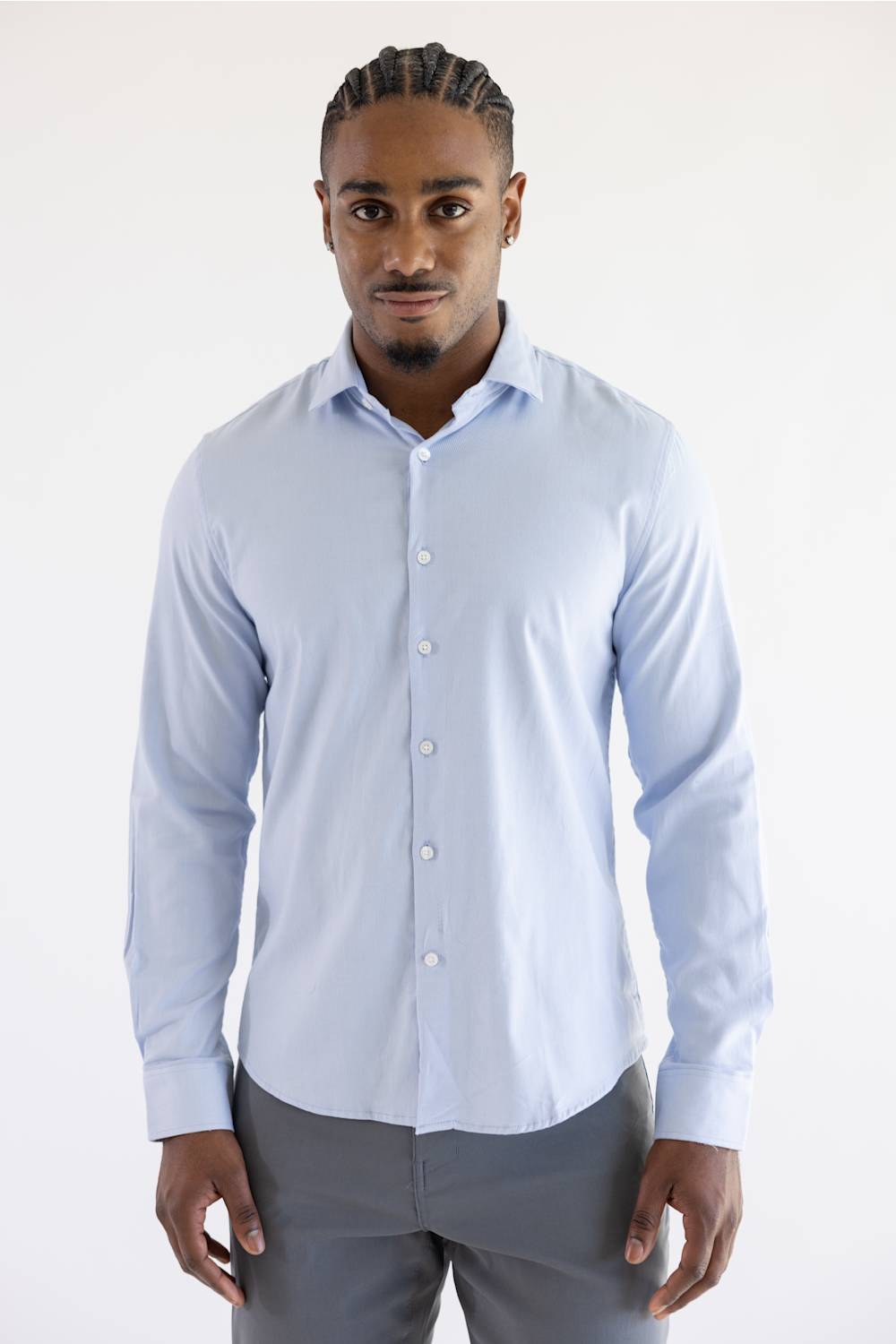 A man wearing a light blue long-sleeve button-down dress shirt and gray trousers, standing against a plain white studio background.