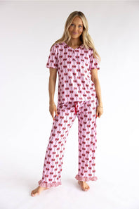 A woman modeling a two-piece pink pajama set with a red bow pattern, consisting of a short-sleeve top and matching pants, against a plain white studio background.