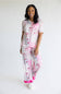 A woman modeling a matching two-piece pajama set with a white base and pink floral pattern, featuring a short-sleeve top and matching pants with bright pink ruffle trim at the cuffs, photographed against a plain white studio background.