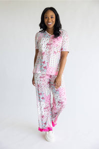 A woman modeling a matching two-piece pajama set with a white base and pink floral pattern, featuring a short-sleeve top and matching pants with bright pink ruffle trim at the cuffs, photographed against a plain white studio background.