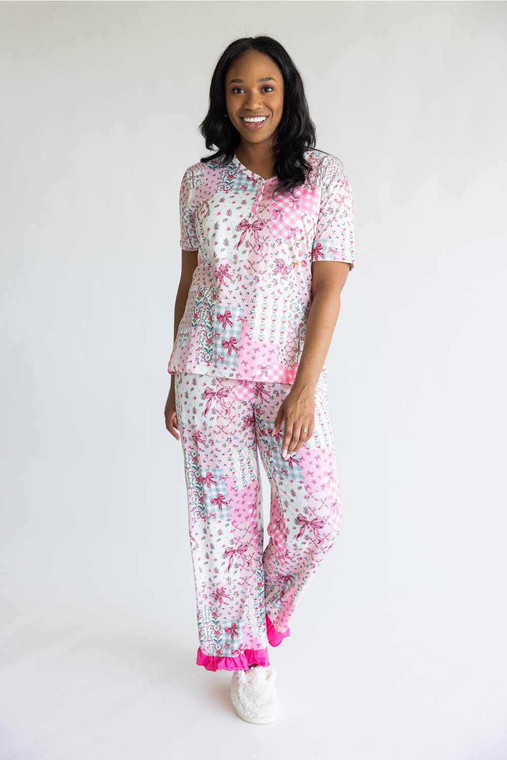 A woman modeling a matching two-piece pajama set with a white base and pink floral pattern, featuring a short-sleeve top and matching pants with bright pink ruffle trim at the cuffs, photographed against a plain white studio background.