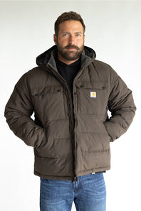 A man wearing a brown Carhartt hooded puffer jacket over a black shirt and blue jeans, standing against a light gray studio background.