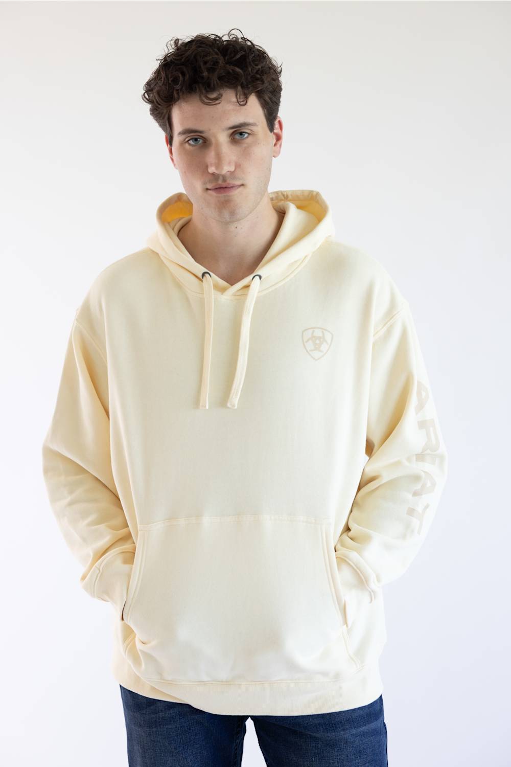 A man wearing a cream-colored hooded sweatshirt with a drawstring and a small shield logo on the left chest, paired with dark blue jeans, photographed against a plain white studio background.